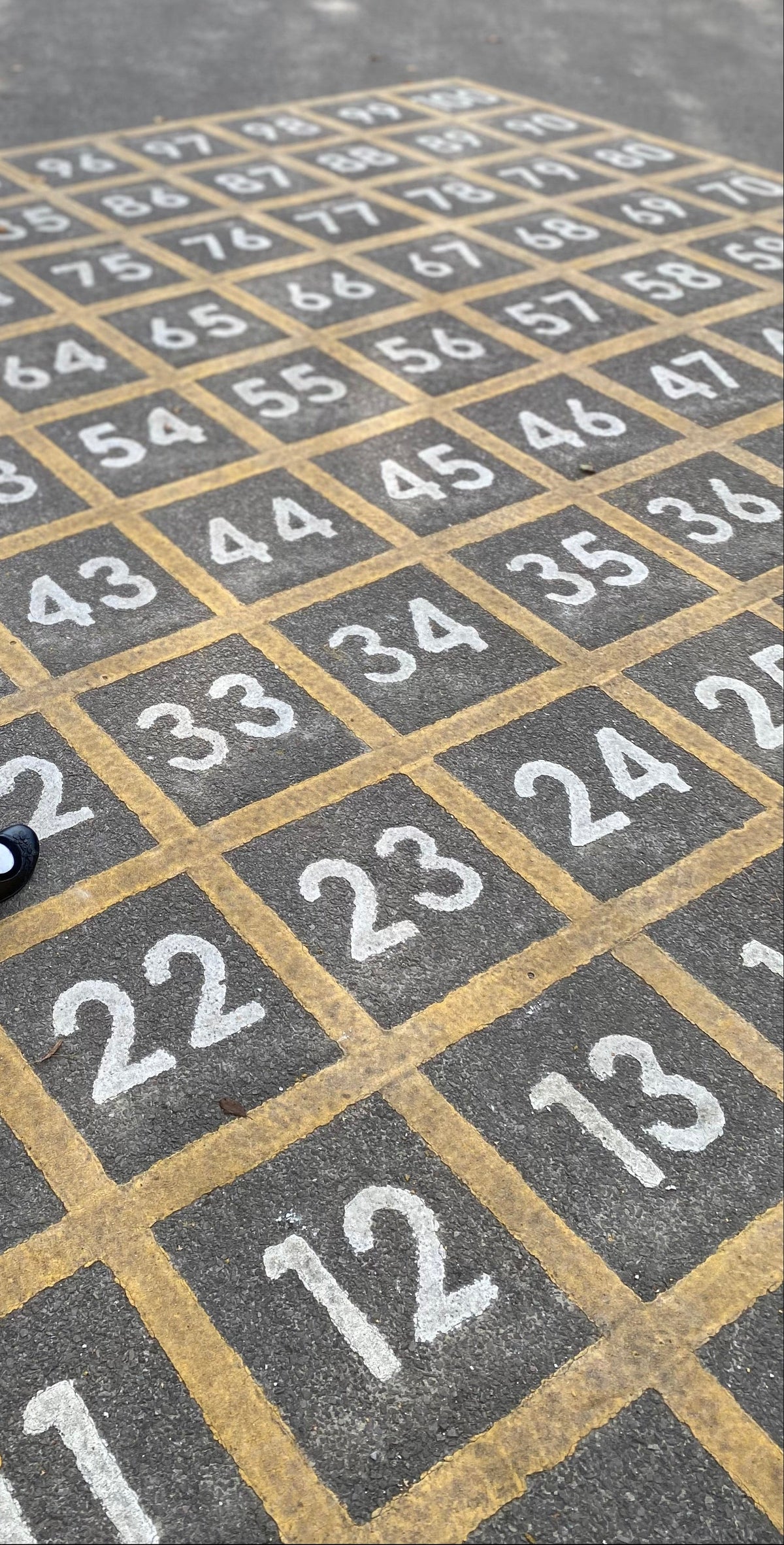 A young girl in a school uniform walks across a large outdoor hopscotch court with numbered squares painted on the pavement.
